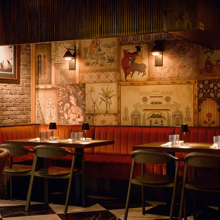 Cozy restaurant interior with red cushioned bench seating, wooden tables and chairs, wall art depicting traditional Indian scenes, and warm ambient lighting.